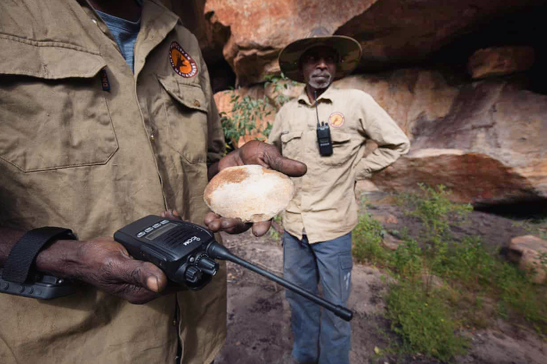 Indigenous ranger Berribob Watson holds modern and ancient technology, a two-way radio and a stone used for grinding pigments for painting. Warddeken ranger Ricky Nabarlambarl stands behind